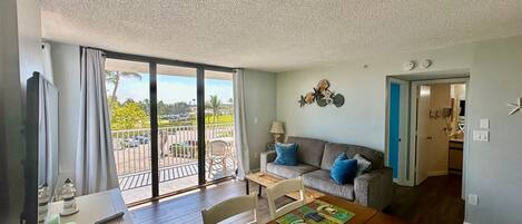 A living room with a sofa, a dining table with placemats, a TV, and a balcony with a view of a parking lot and palm trees.