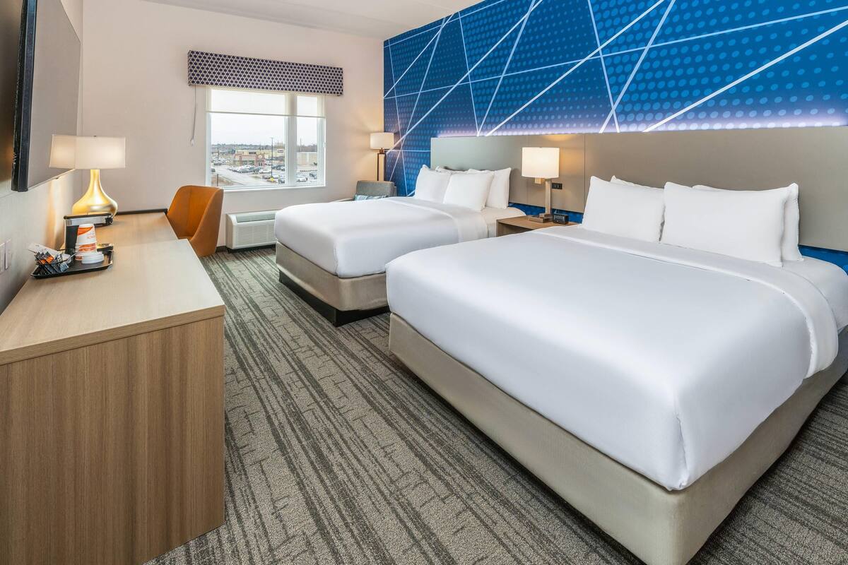Habitación de hotel con dos camas, un escritorio, una silla y un ventanal grande.