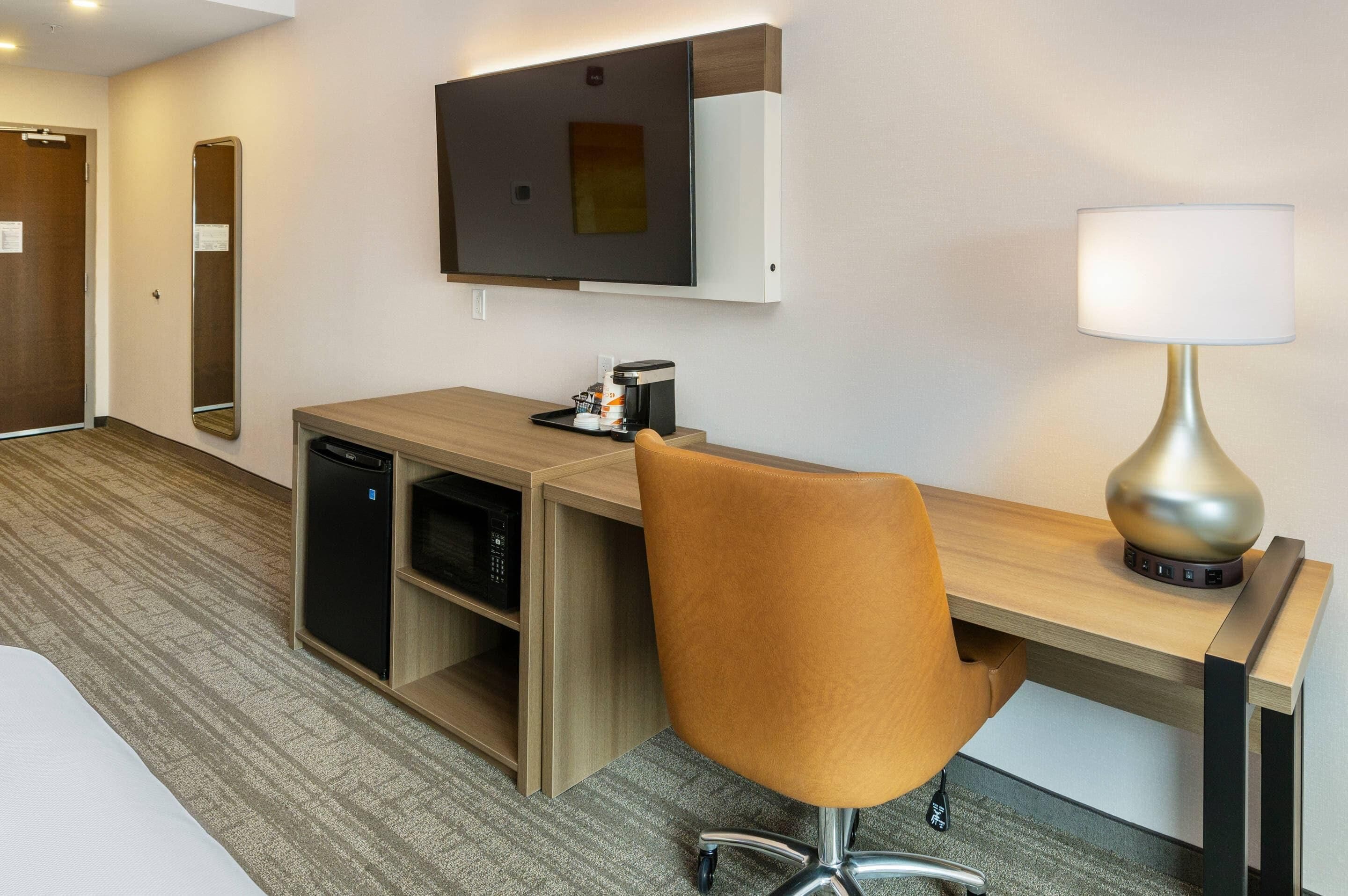 Ein Hotelzimmer mit Schreibtisch aus Holz, einem Stuhl, einer Lampe, einer Kaffeemaschine, einem kleinen Kühlschrank und einem Fernseher.