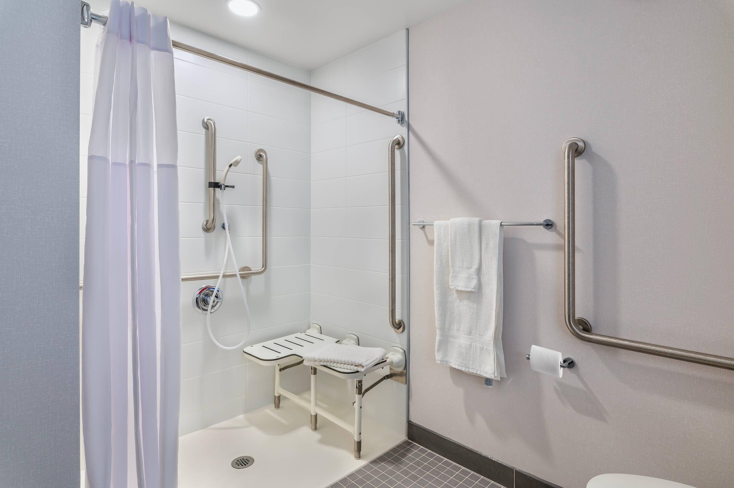 Ein Badezimmer mit Dusche, Haltegriffen, einer rollstuhlgerechten Sitzbank und einem WC mit Haltegriff.