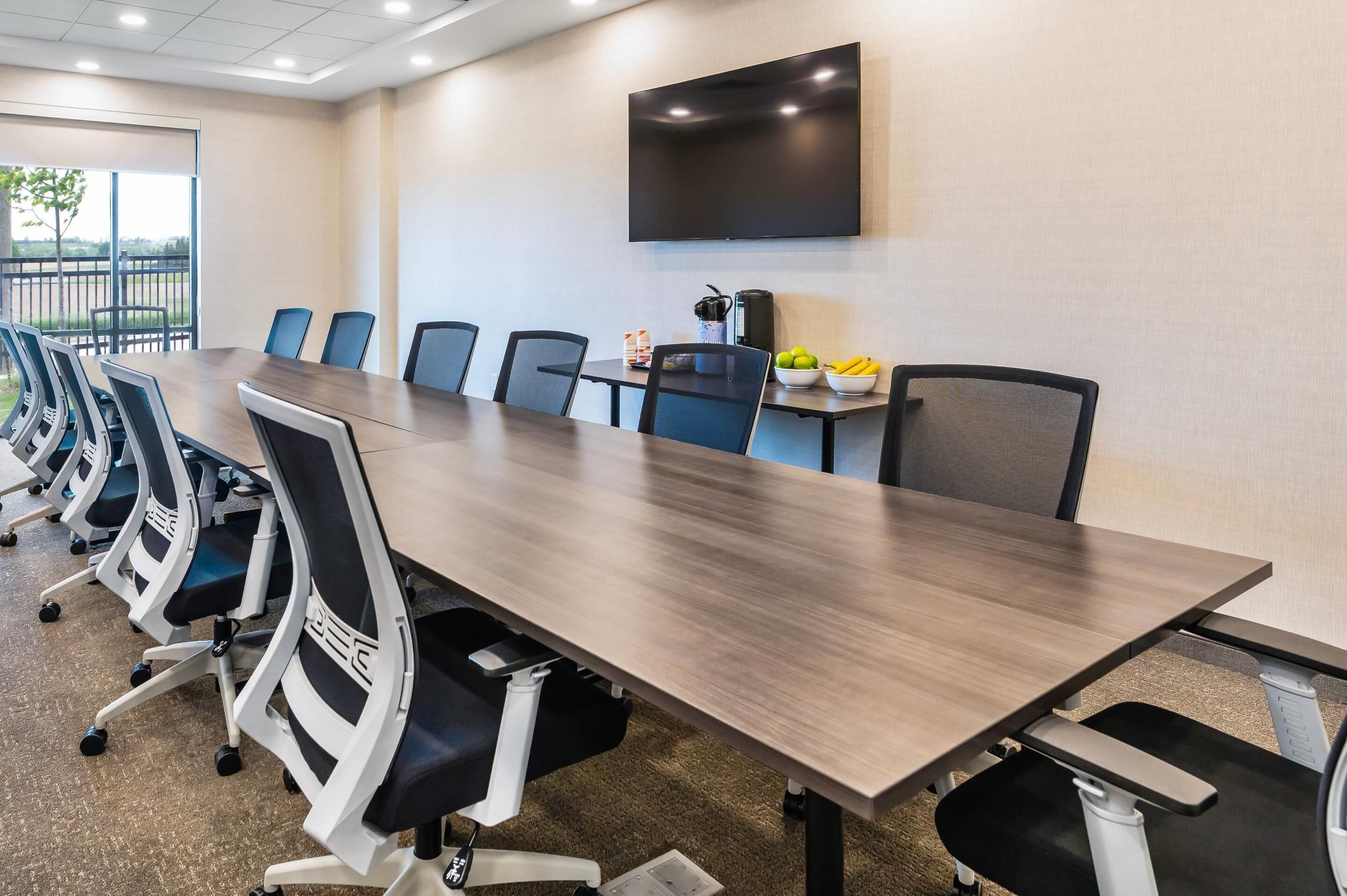 A conference room with a long table, multiple chairs, a flat-screen TV, and a small table with fruits and a coffee maker.