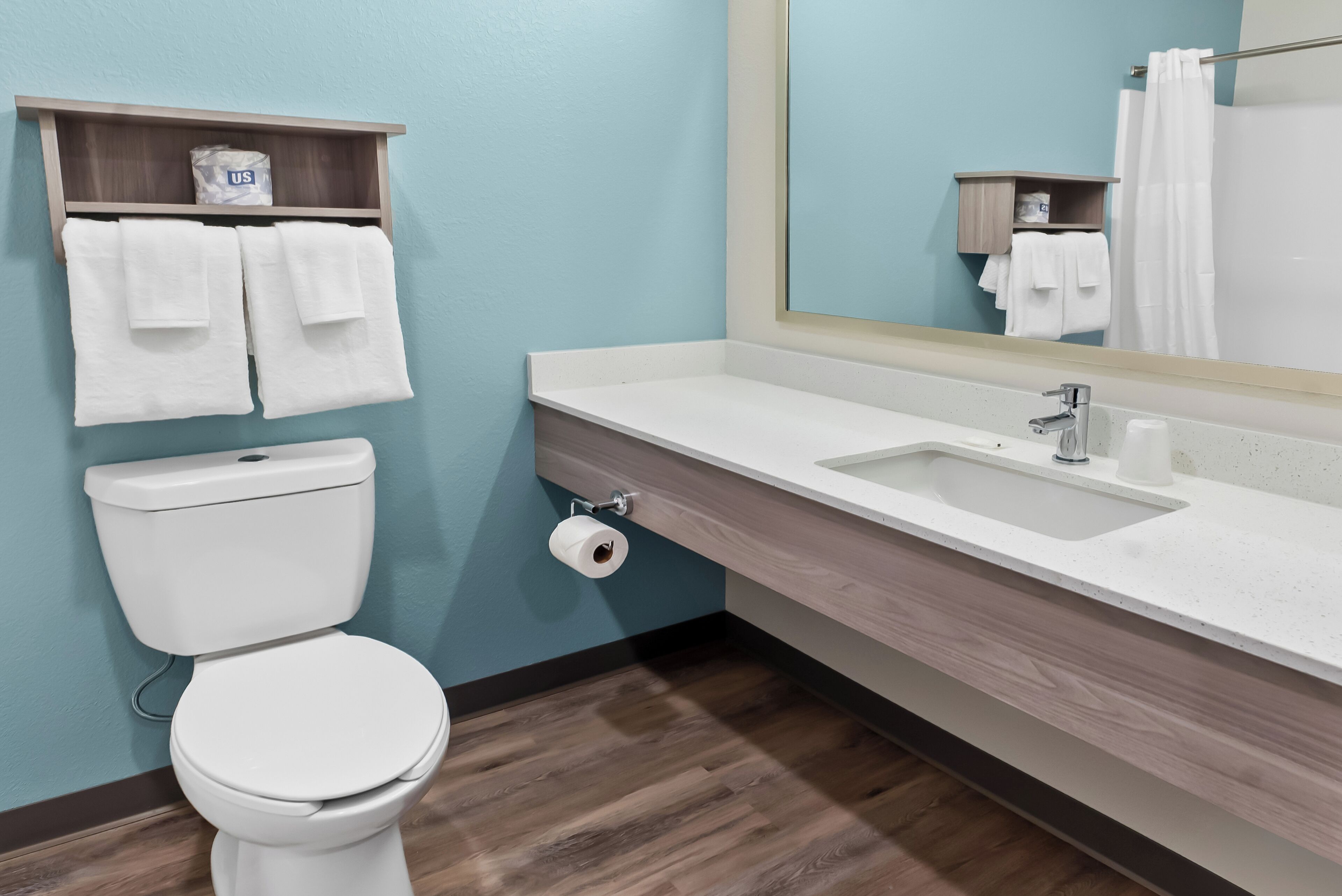 A bathroom with a toilet, sink, and mirror. There are towels hanging on the wall and a shelf with toiletries.