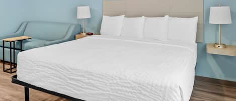 Una cama bien hecha con colcha blanca, un sillón azul claro y dos mesitas de noche con lámparas.