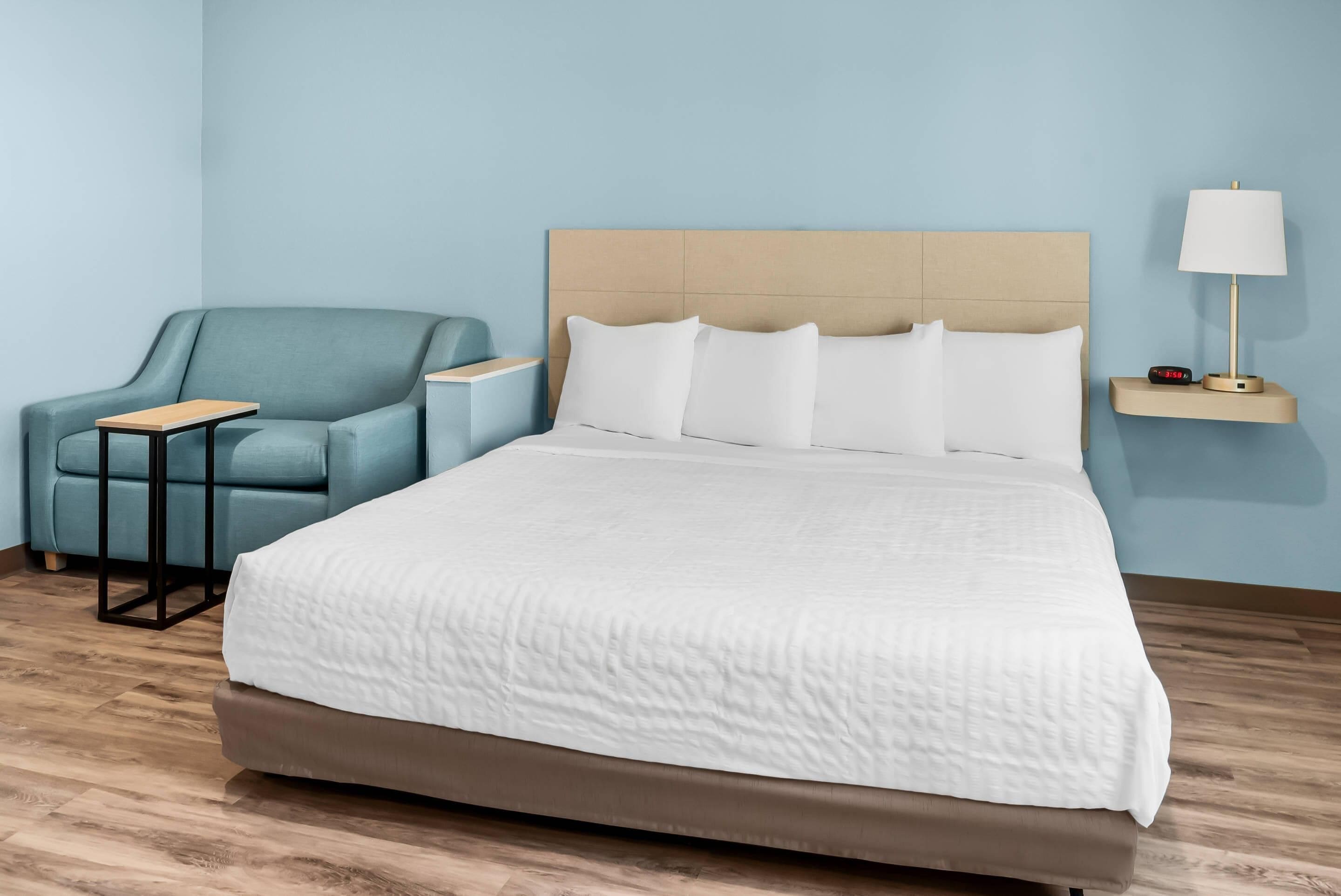 A hotel room with a large bed, a blue armchair, a small table, and a bedside lamp.