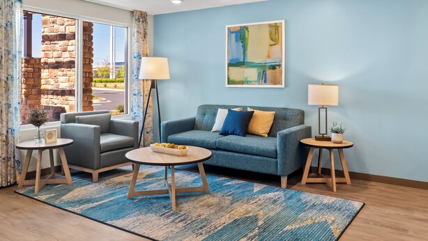 A modern living room with a blue sofa, two side tables, a coffee table, and a large window with a view of a stone building. - Woodspring Suites Greenville Airport (Greenville)