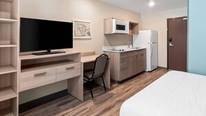 A compact hotel room with a bed, a desk with a flat-screen TV, a chair, a kitchenette with a microwave and refrigerator, and a wooden floor. - Woodspring Suites Greenville Airport (Greenville)