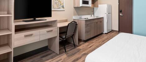 A compact hotel room with a bed, a desk with a flat-screen TV, a chair, a kitchenette with a microwave and refrigerator, and a wooden floor.