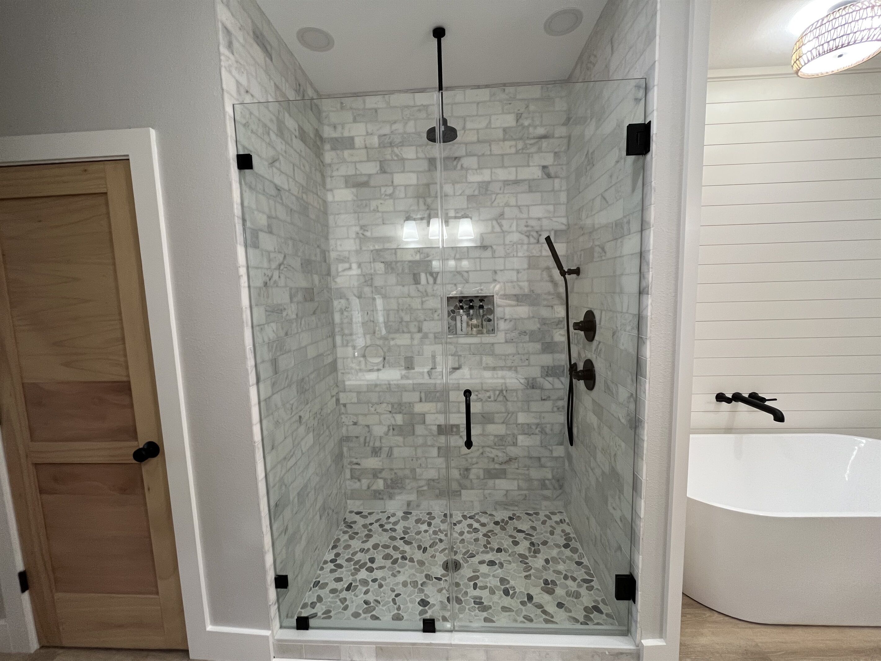 A glass-enclosed shower with a pebble-patterned floor, a white bathtub, and a wooden door.
