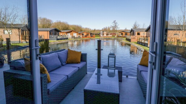 A modern outdoor seating area with a glass barrier, a water canal, and wooden buildings in the background. - Coniston View Lodge (Carnforth)