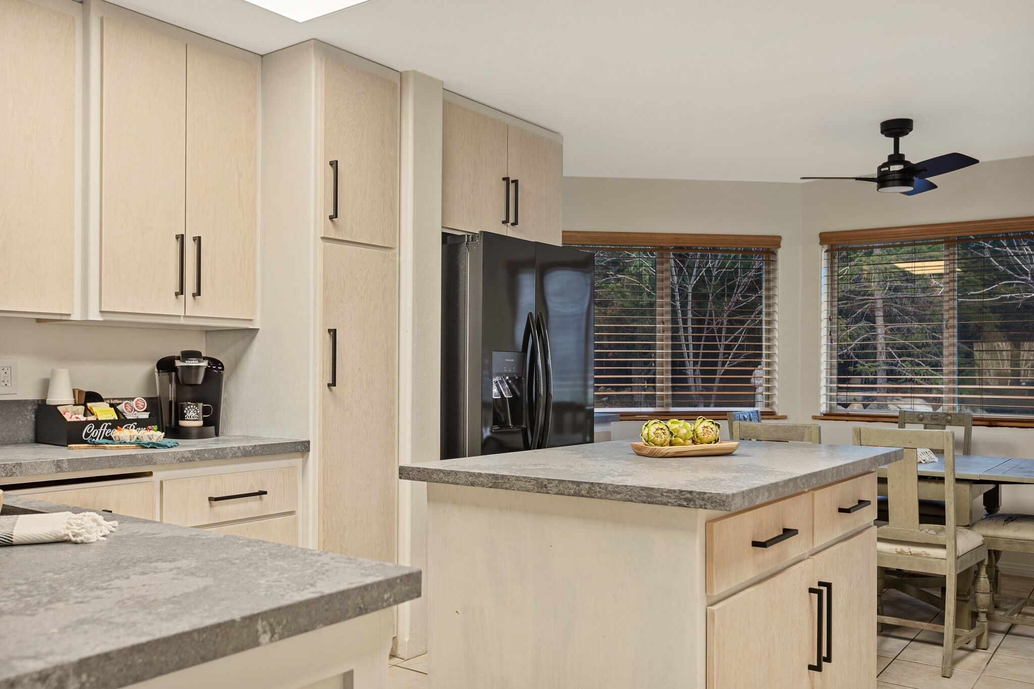 A modern kitchen with light wood cabinets, a stainless steel refrigerator, and a granite countertop.