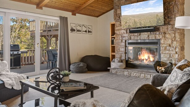 Una sala de estar acogedora con chimenea de piedra, un televisor de pantalla plana montado en una pared de piedra y un ventanal corredera que da a una terraza exterior con una parrilla.