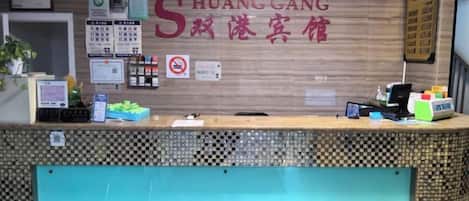 A hotel reception desk with a check-in counter, a wall-mounted sign with hotel name, a no-smoking sign, and various informational posters.