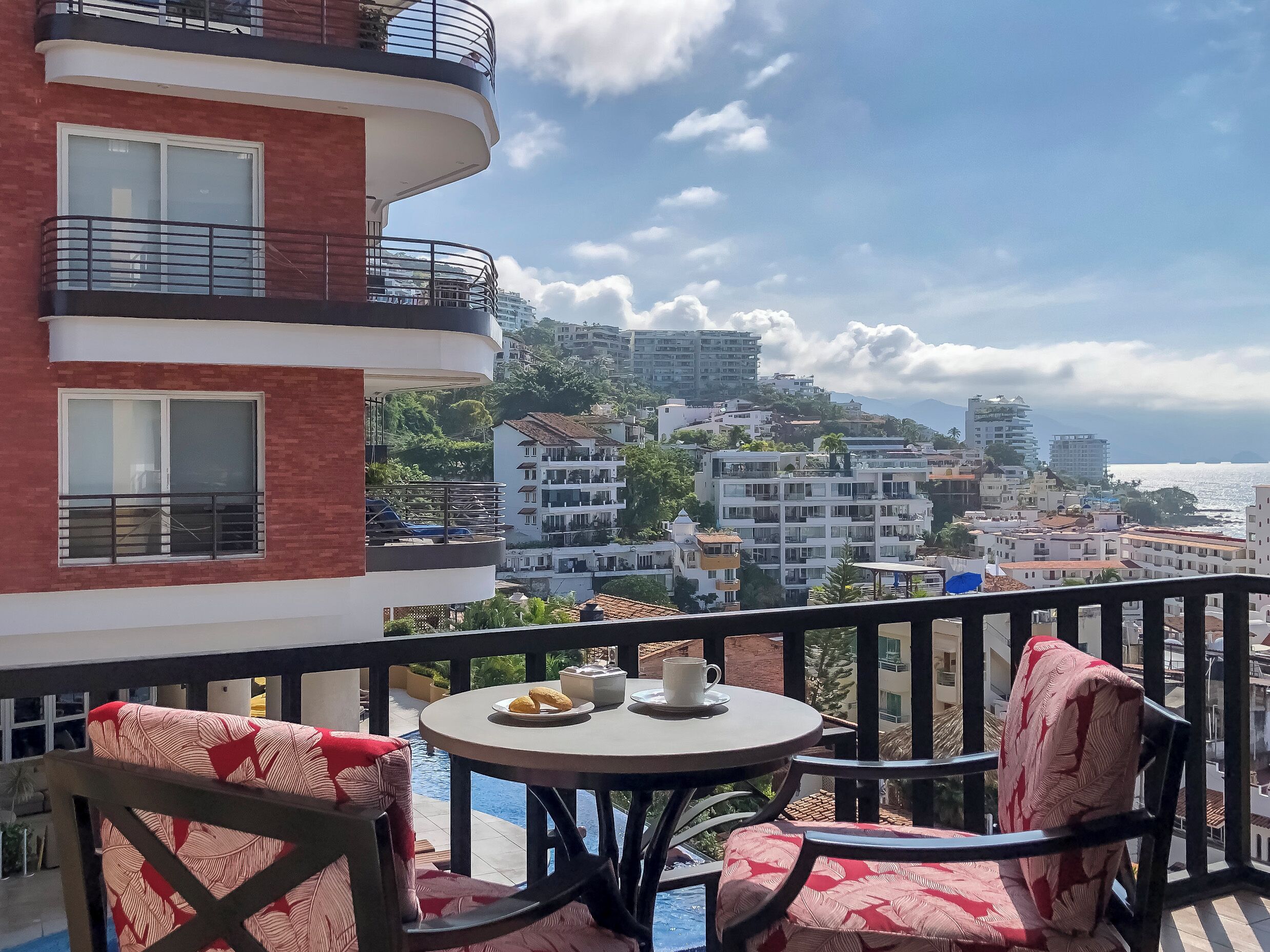 A balcony with a table and chairs, overlooking a cityscape with buildings and a body of water.