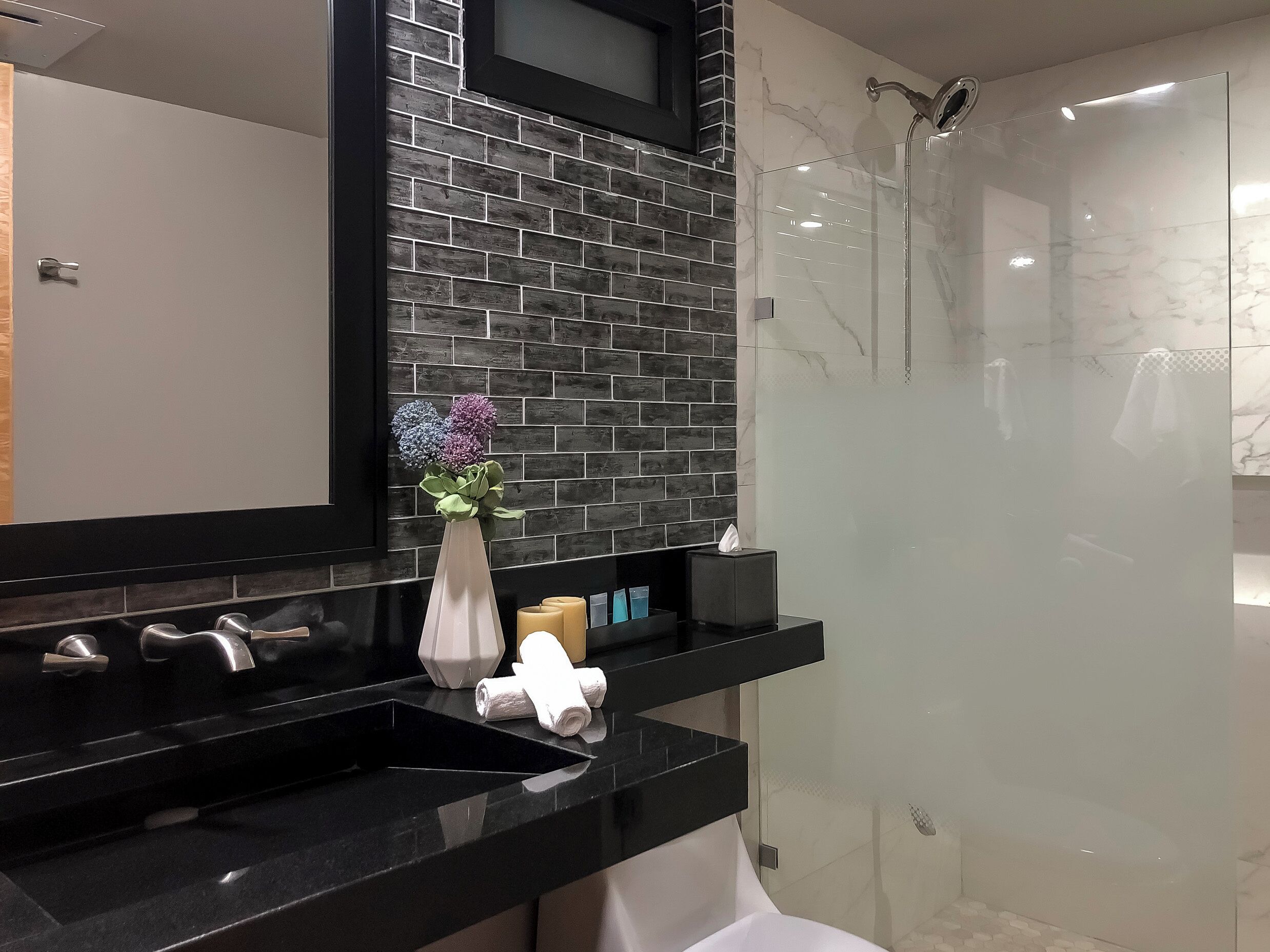 Une salle de bain moderne avec un plan de toilette noir, un miroir, un vase de fleurs et une cabine de douche en verre.