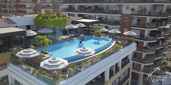 A rooftop pool with sun loungers and umbrellas, surrounded by balconies and greenery.
