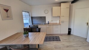 A compact kitchen with a sink, stove, and cabinets. There is a dining table with chairs and a small potted plant. A clock is on the wall, and a door is visible. - Vacation apartment / fitter's apartment / short-term rental (Schönborn)