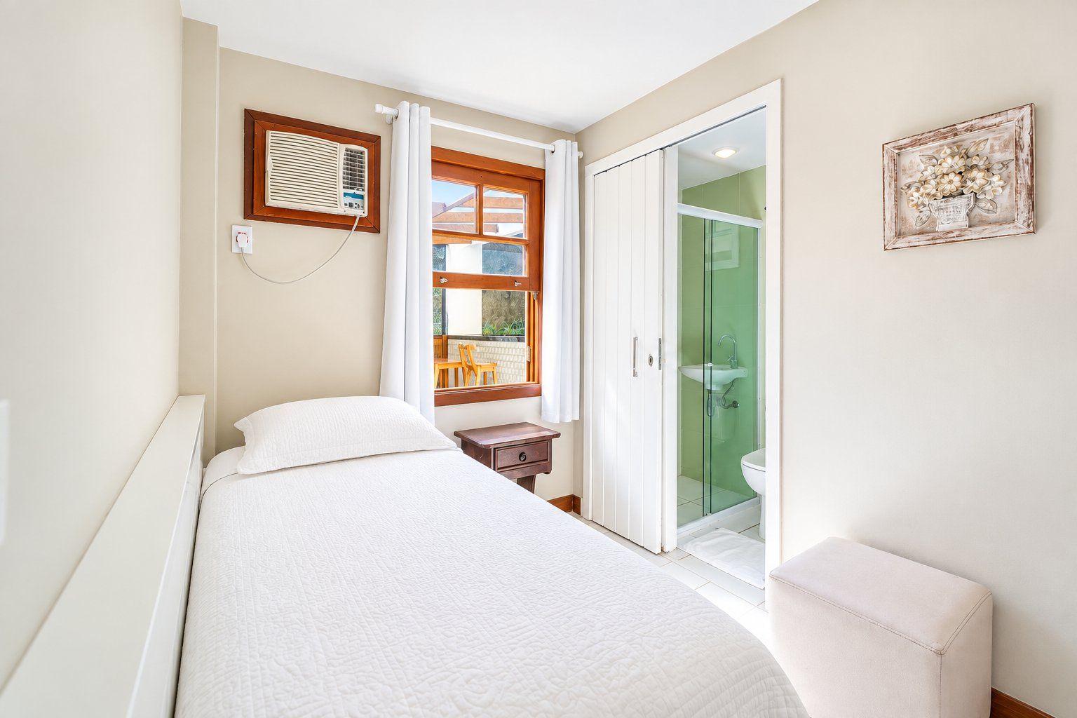 A bedroom with a bed, a bedside table, a window with curtains, a radiator, a wall-mounted air conditioner, a glass shower enclosure, and a framed picture on the wall.