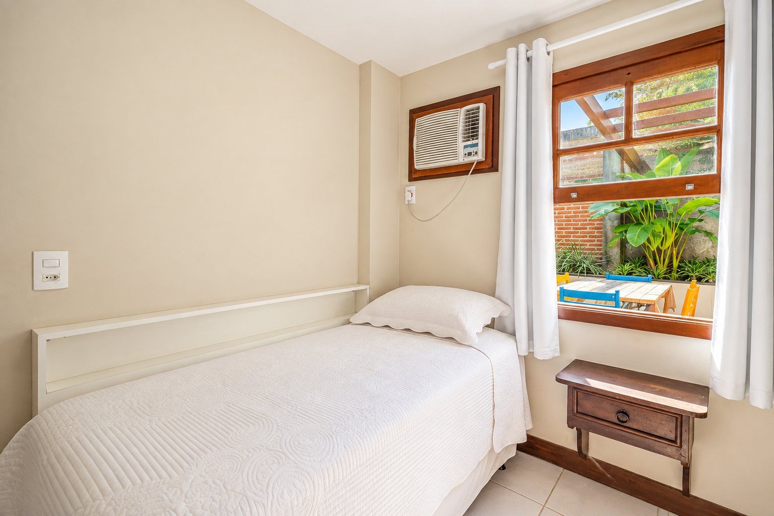 A bedroom with a bed, a bedside table, a window with curtains, and a wall-mounted air conditioning unit.