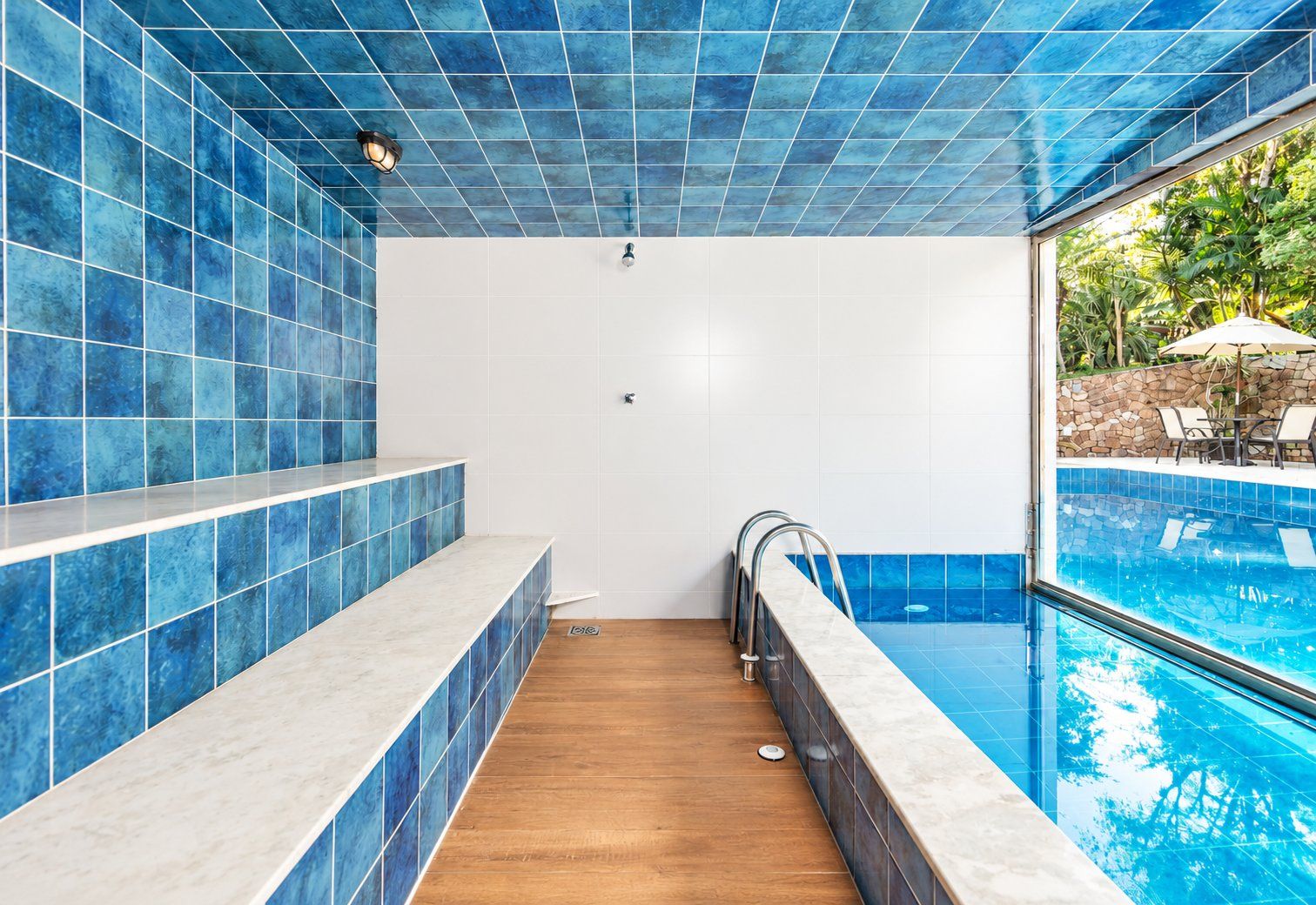 Uma área de piscina com azulejos azuis, piso de madeira e um espaço de estar com cadeiras e um guarda-sol.