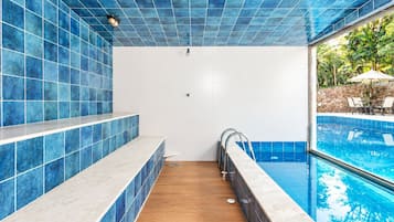 Uma área de piscina com azulejos azuis, piso de madeira e um espaço de estar com cadeiras e um guarda-sol.