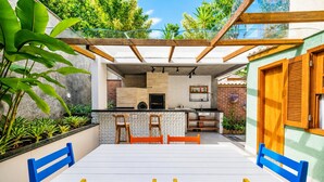 A patio with a dining table and chairs, a kitchen area with a counter and stools, and a view of the surrounding greenery. - Bz21 Casa com Jacuzzi e churrasqueira (Buzios)