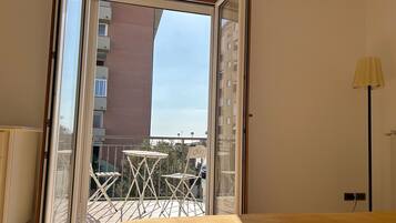 A room with a bed, a window with a view of a building, and a balcony with a table and chairs.