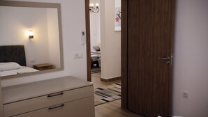 Quarto de hotel com uma cama, penteadeira, espelho, luminária na parede e uma porta que dá para outro cômodo.