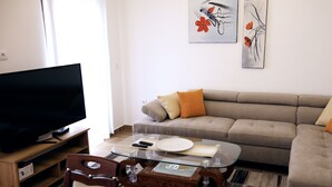 Uma sala de estar com televisão de tela plana, mesa de centro de vidro, sofá bege com almofadas laranja e duas obras de arte penduradas na parede.