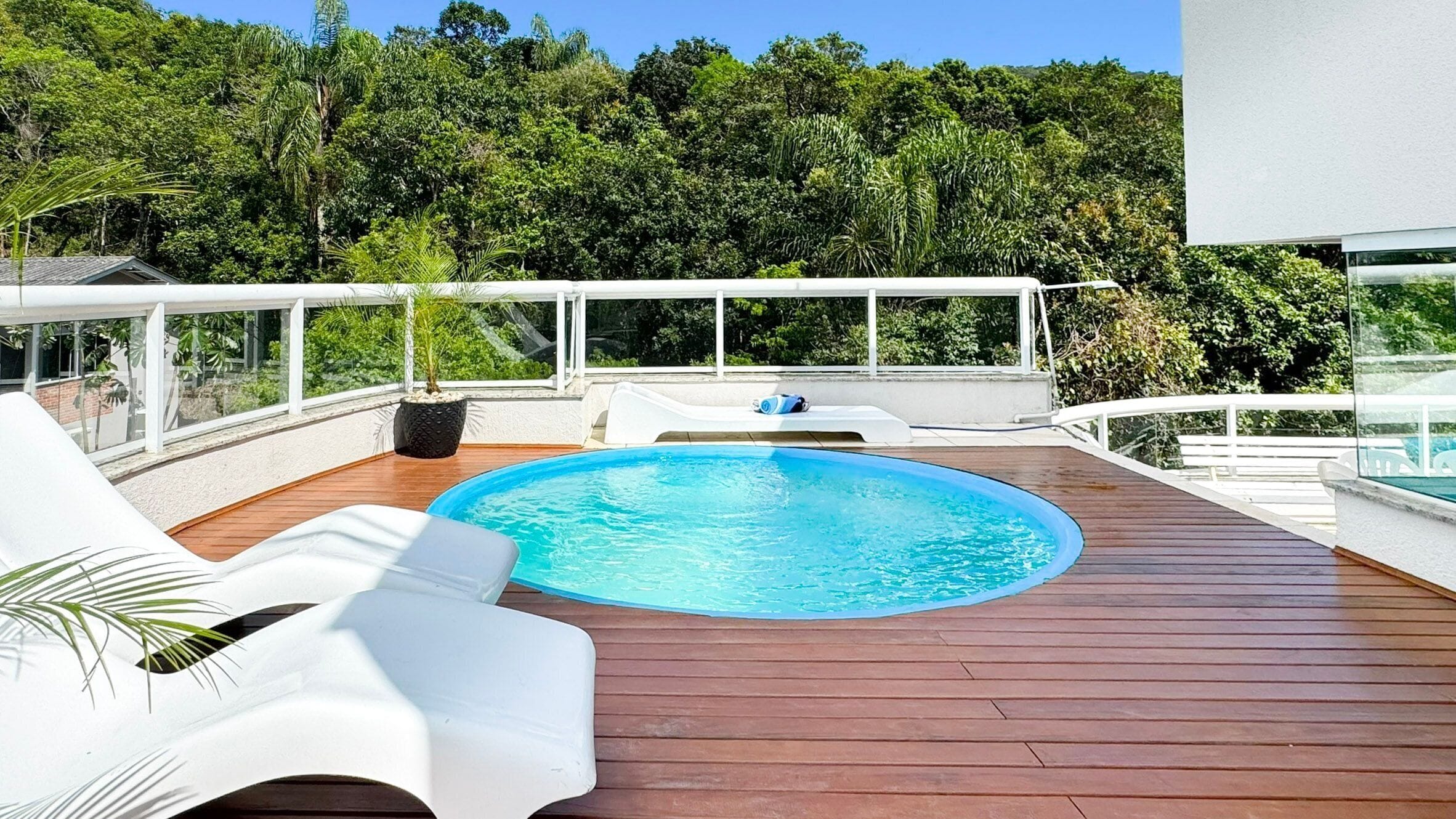 A rooftop pool with wooden decking, white lounge chairs, and a view of dense greenery in the background.