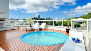 A rooftop pool with wooden decking, two white lounge chairs, and a view of the cityscape. - Cobertura com Piscina na praia de Bombas (Bombinhas)