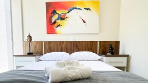 Ein Hotelzimmer mit hölzernem Kopfteil, einem Bett mit weißer Bettwäsche, einem farbenfrohen abstrakten Gemälde an der Wand und einer Buddha-Statue auf dem Nachttisch.