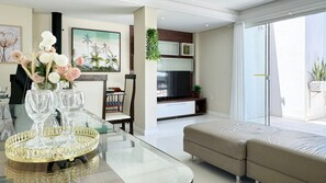 A modern living room with a glass coffee table, a beige sofa, a TV, and a dining area with chairs and a table. - Cobertura com Piscina na praia de Bombas (Bombinhas)