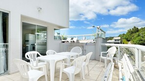 A modern outdoor patio with white plastic chairs and tables, a glass door leading to a kitchen, and a view of a residential area. - Cobertura com Piscina na praia de Bombas (Bombinhas)