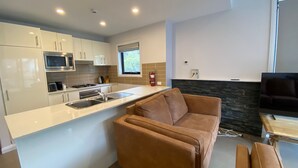 A modern kitchen with white cabinets, a stainless steel microwave, and a gas stove. There is a brown leather sofa and a television in the living area.