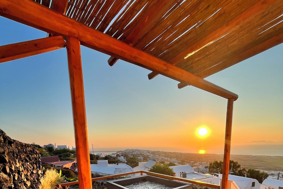 A sunlit outdoor seating area with a stone wall and cactus, overlooking a coastal town at sunset.