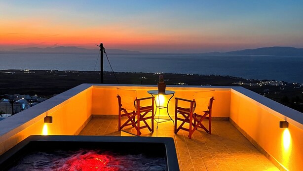 A rooftop terrace with a hot tub, two chairs, and a table under a sunset sky. - Sunset Paradise Oia (Santorini)