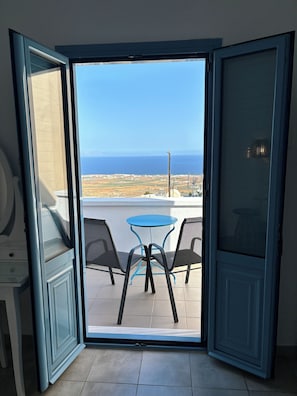 An open sliding glass door leading to a balcony with a table and chairs, overlooking a coastal landscape.