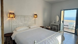 A bedroom with a white metal bed, two wall-mounted lamps, a small bedside table, and a view through a glass door to an outdoor area. - Sunset Paradise Oia (Santorini)
