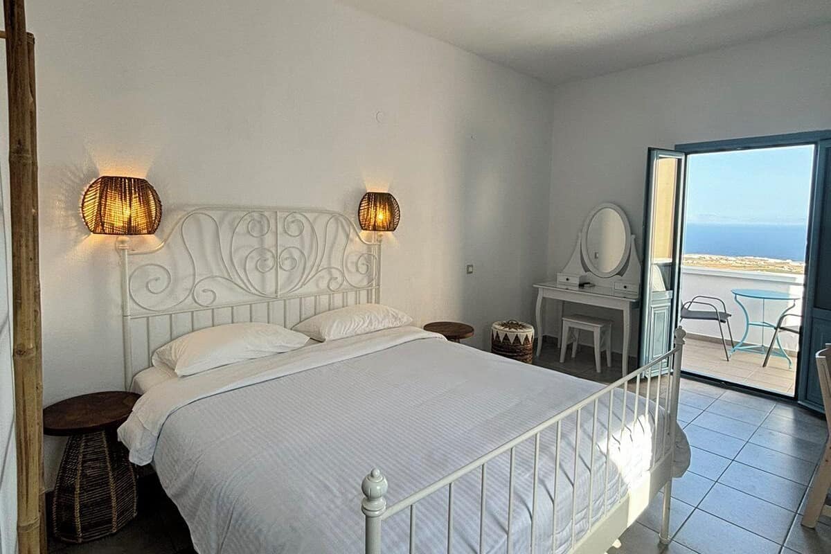 A bedroom with a white metal bed, two wall-mounted lamps, a small bedside table, and a view through a glass door to an outdoor area.