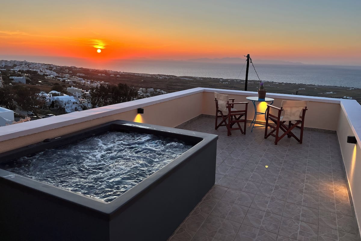A rooftop terrace with a hot tub, a table with chairs, and a view of the sunset over the sea.