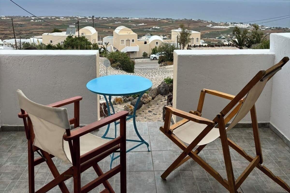 A balcony with two wooden chairs and a small round table, overlooking a coastal village with white buildings and a view of the sea and mountains.
