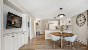 A modern kitchen with white cabinetry, a wooden dining table with chairs, a large clock on the wall, and a flat-screen TV mounted on the wall. - Satsuma | Close to Waterpark, Hot Springs & Golf (Fairmont Hot Springs)