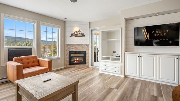 A spacious living room with a fireplace, a large window, a TV, and a ceiling fan. - Satsuma | Close to Waterpark, Hot Springs & Golf (Fairmont Hot Springs)