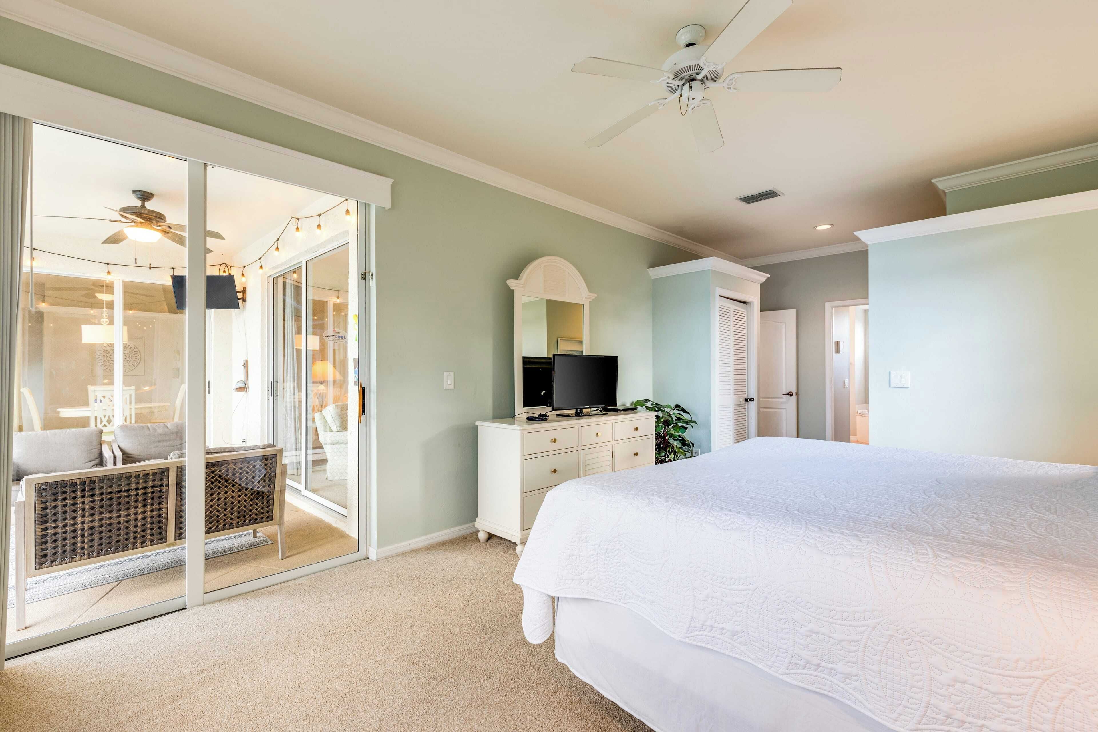 A bedroom with a large bed, a TV, a dresser, and a sliding glass door leading to a balcony with outdoor seating.