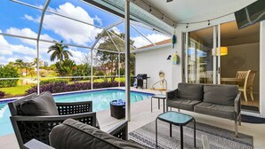 A poolside area with a glass-enclosed patio, a gray sofa, a small table, and a chair. - Private Pool, Pond Views, & Bonus Lounge in Gated Naples Community Drive to Dining & Sand (Naples)