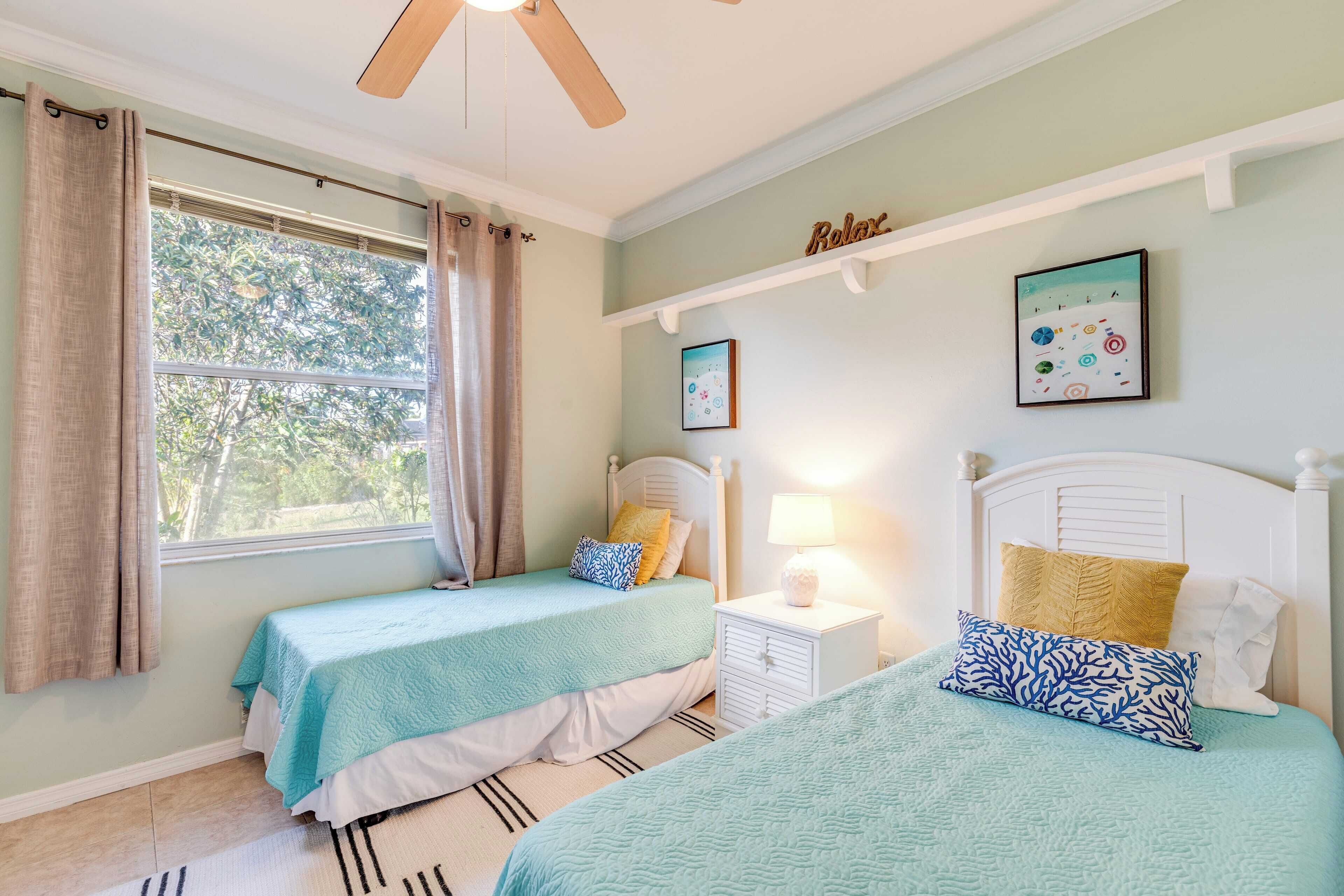 Een slaapkamer met twee bedden, een raam met gordijnen, een plafondventilator en een wandplank met foto’s.