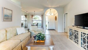 A living room with a beige sofa, a coffee table with a floral arrangement, a TV on a cabinet, and a dining area in the background. - Private Pool, Pond Views, & Bonus Lounge in Gated Naples Community Drive to Dining & Sand (Naples)