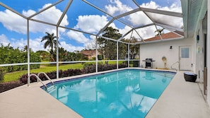 Une piscine couverte avec vue sur une pelouse et des arbres, un barbecue et un tuyau d’arrosage.