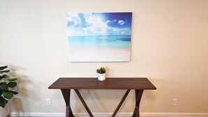 A wooden table with a plant on top, positioned against a wall with a painting of a beach scene. - The Austin Vacation (Austin)