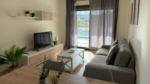 Ein modernes Wohnzimmer mit Flachbildfernseher auf einer Holzkonsole, einem grauen Sofa, einem Couchtisch und einem Esstisch mit zwei dekorativen Vasen.